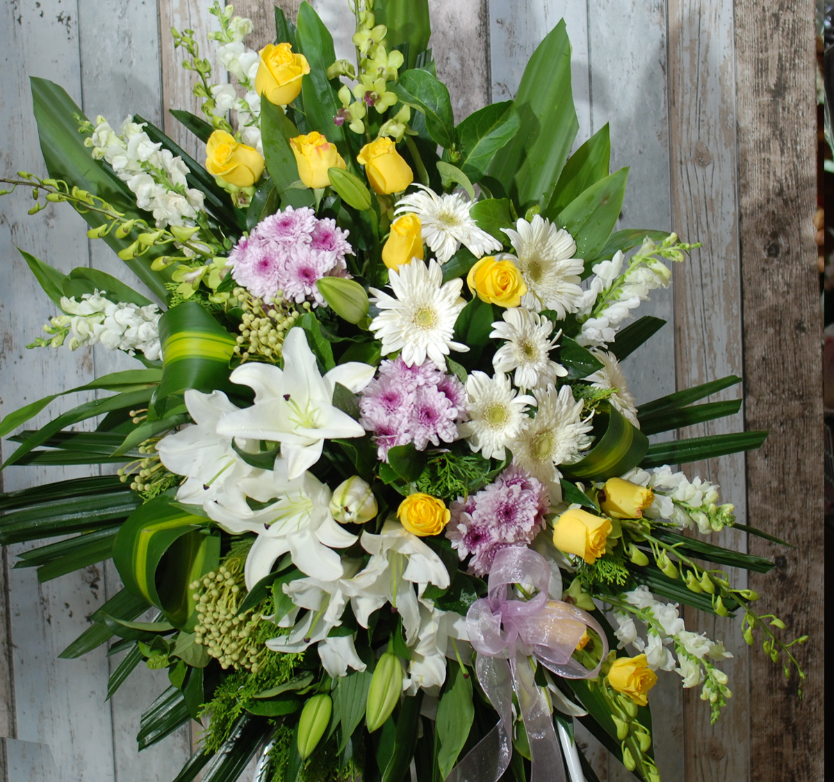 A floral arrangement with yellow roses, white lilies, white daisies, and pink carnation accents amidst green foliage, displayed against a textured wooden backdrop.