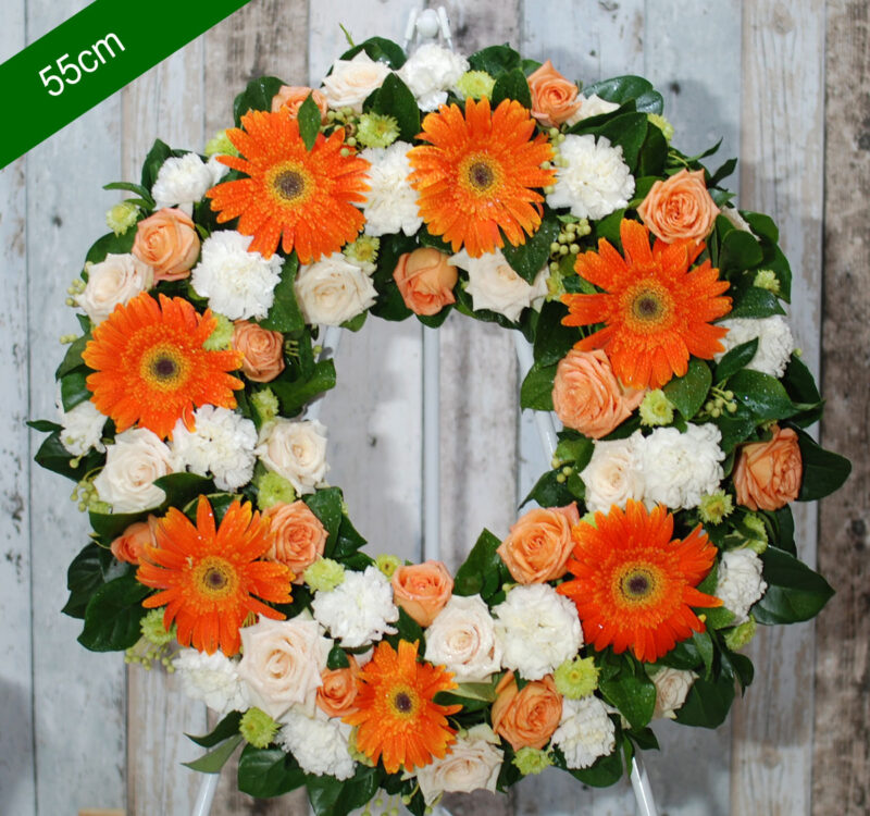 A 55cm floral wreath predominantly featuring orange gerbera daisies, cream roses, and white carnations, with green foliage against a light wooden backdrop.