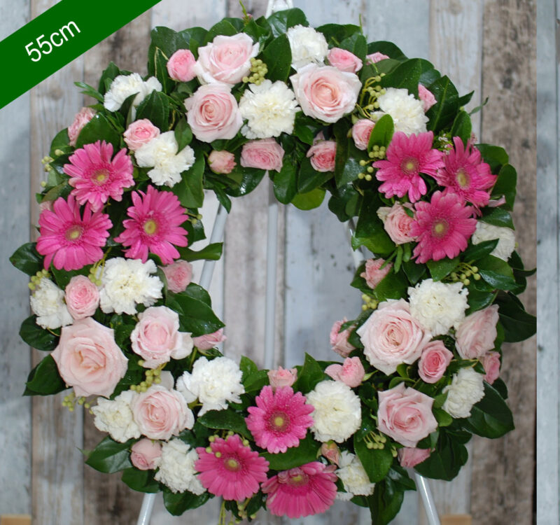 A 55cm floral wreath featuring pink gerbera daisies, pink roses, and white carnations, complemented by green foliage, displayed against a wooden background.