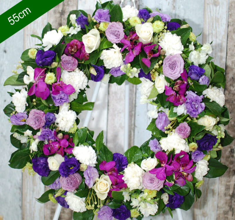 A 55 cm floral wreath featuring a mix of white, purple, and pink flowers with green leaves, displayed on a white metal stand.