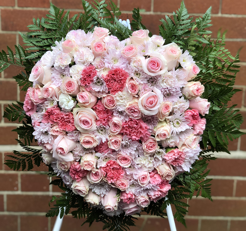A heart-shaped floral arrangement with pink and white roses, pink carnations, and white chrysanthemums, framed by lush green fern leaves, set against a brick wall background.