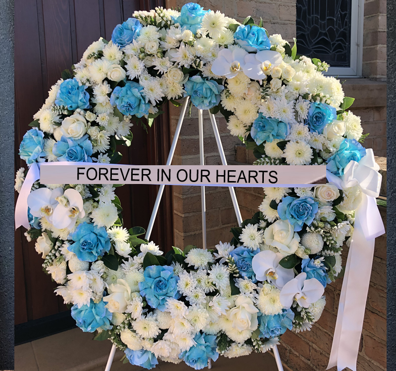 A large floral wreath with blue and white flowers is displayed on a stand in front of a building. A ribbon across the wreath reads "FOREVER IN OUR HEARTS.