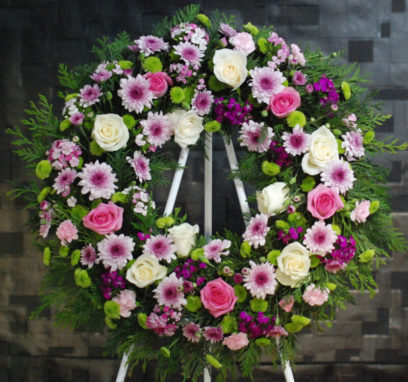 A large floral wreath made of pink, white, and purple flowers with green foliage, displayed on a stand against a dark background.