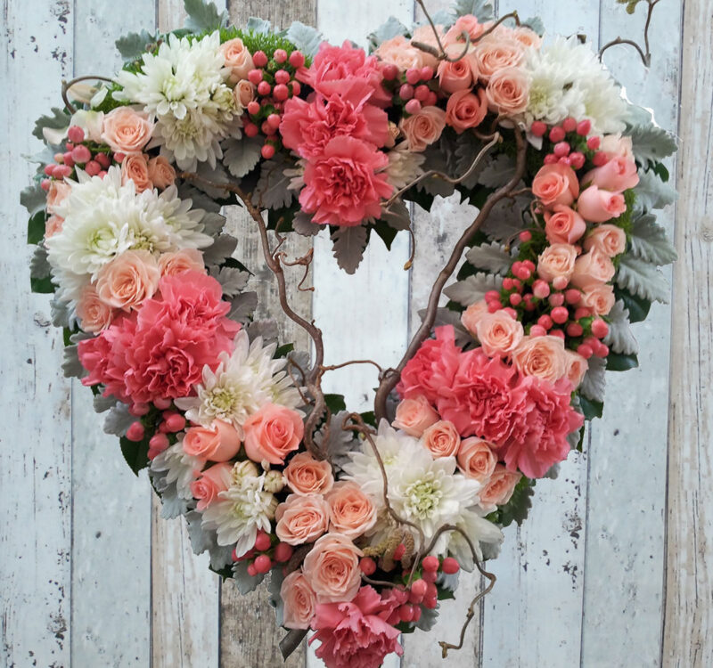 A heart-shaped floral wreath composed of pink and white roses, chrysanthemums, carnations, and assorted greenery, displayed against a rustic wooden background.