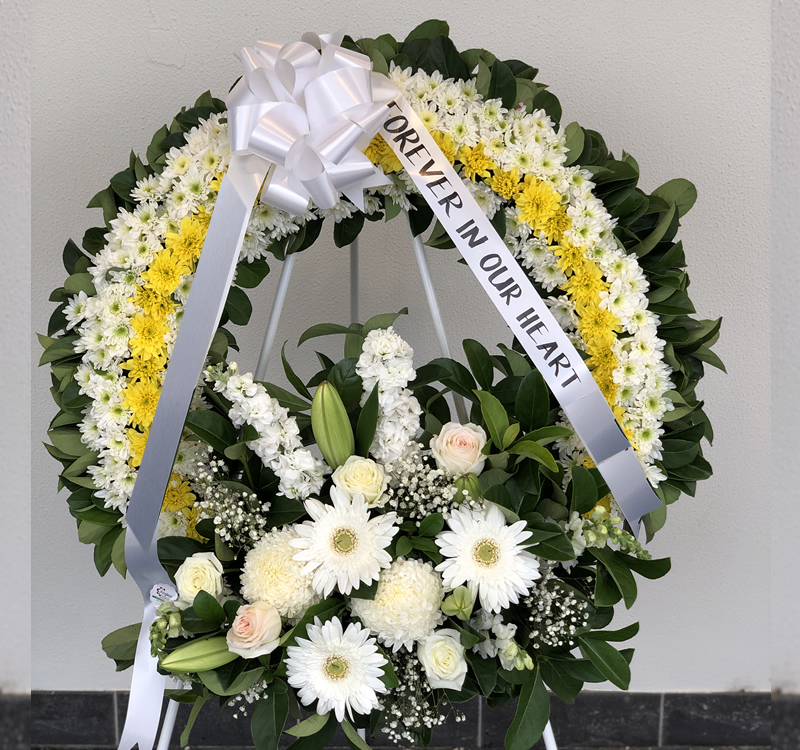 A wreath of white and yellow flowers with a white ribbon inscribed "Forever in Our Heart." The arrangement includes white roses, daisies, and greenery.