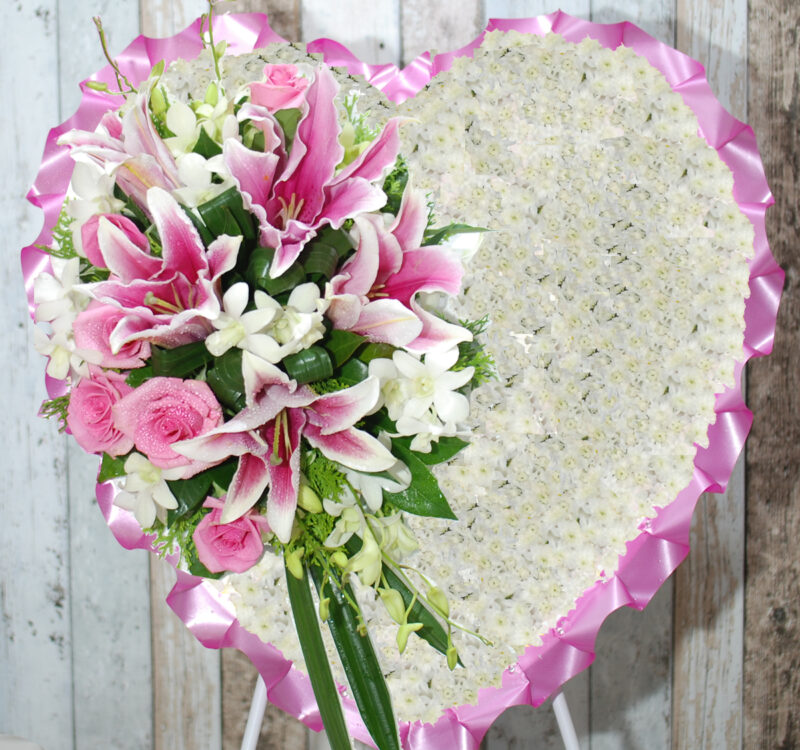 A heart-shaped floral arrangement with a border of pink ribbon features predominantly white flowers and a side bouquet of pink lilies, roses, and white orchids.