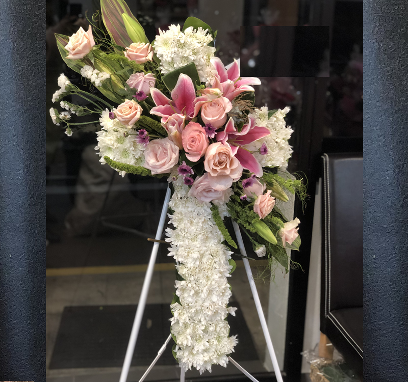 A floral arrangement featuring pink lilies, pale roses, and white flowers on a white tripod stand against a dark background.