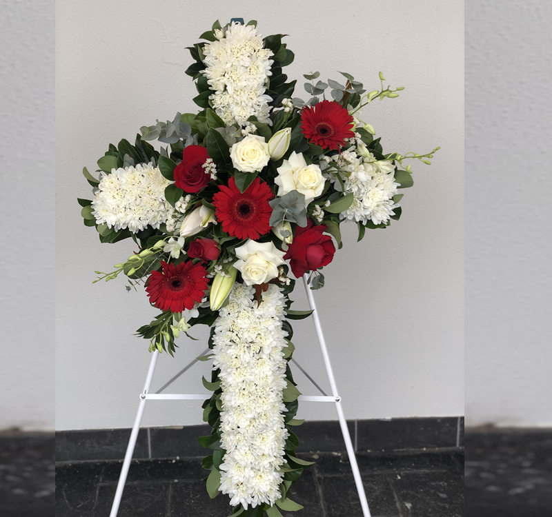 A floral cross arrangement with white chrysanthemums, white lilies, red roses, and red gerberas placed on a white stand against a plain wall.