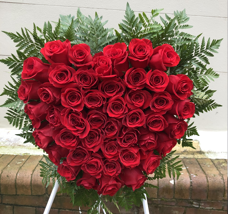 Heart-shaped arrangement of red roses surrounded by green ferns, displayed on a white stand against a brick and concrete background.