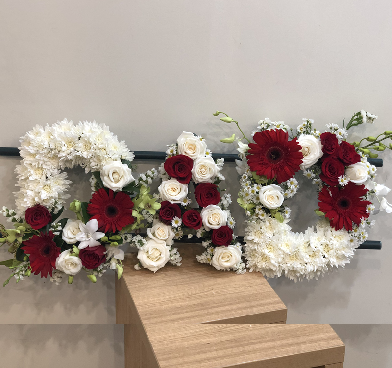 Floral arrangement spelling "DAD" with white and red flowers, displayed on a wooden platform against a light wall.