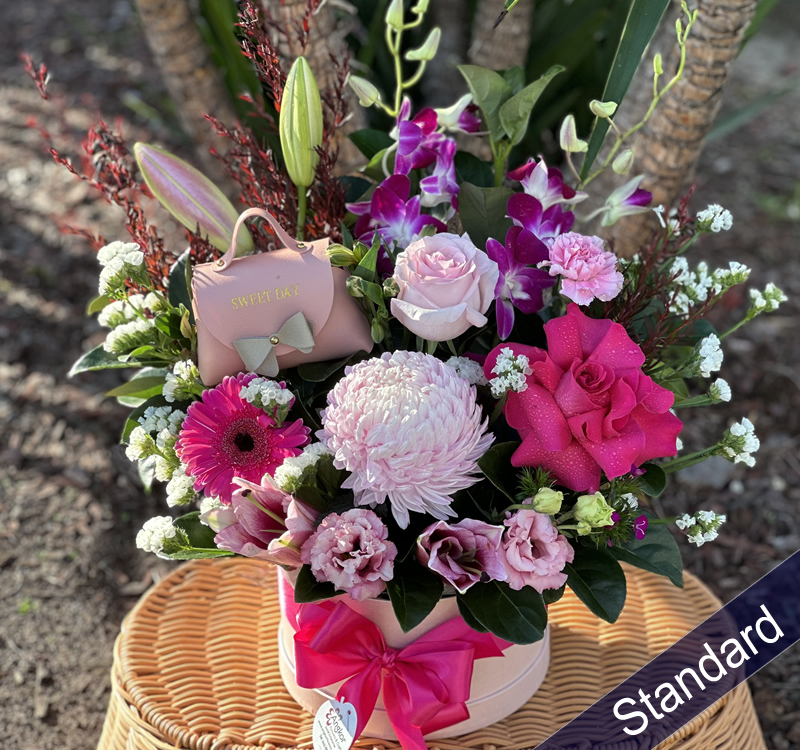 A wicker basket holds a vibrant floral arrangement featuring lilies, roses, carnations, and other mixed flowers with varied colors. A small pink gift bag with "SWEET DAY" is included. Text reads "Standard".