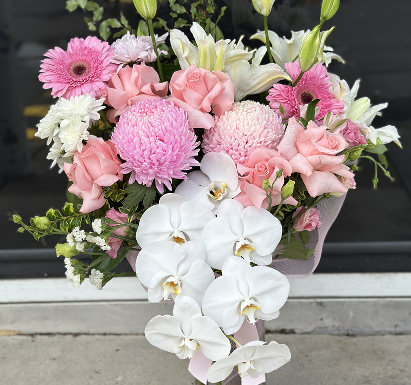 A large bouquet featuring white orchids, pink roses, pink chrysanthemums, gerbera daisies, lilies, and assorted greenery, arranged in front of a dark window.