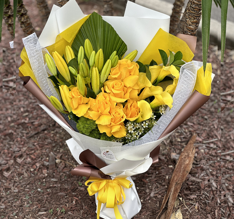 A bouquet of yellow lilies and roses wrapped in white, yellow, and brown paper with a yellow ribbon, placed on mulch near palm tree trunks.