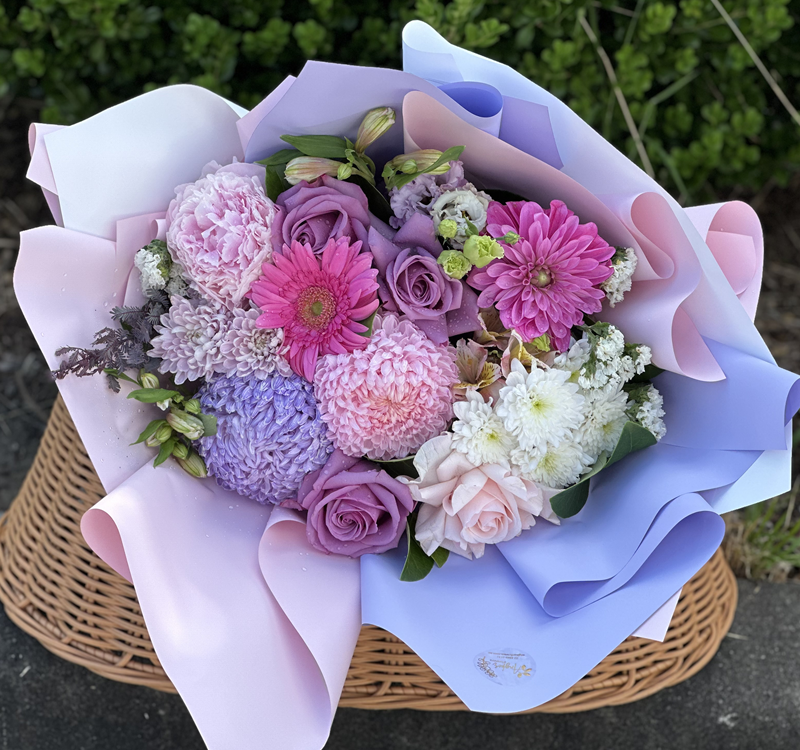A bouquet of pink, purple, and white flowers wrapped in pastel paper, placed on a wicker basket with greenery in the background.