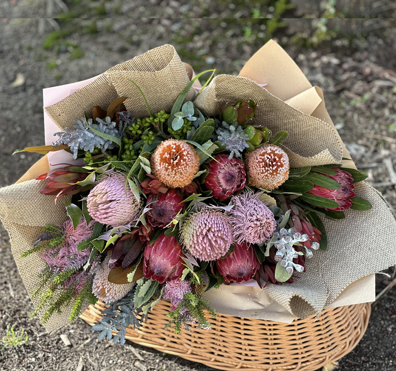 A wicker basket filled with a bouquet of various protea flowers and greenery, wrapped in burlap and paper, placed outdoors on the ground.